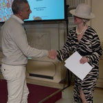 Two people shaking hands in a formal setting with a screen displaying text in the background.