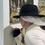 Person wearing a black hat and white outfit with a pearl necklace, looking at text on a wall.