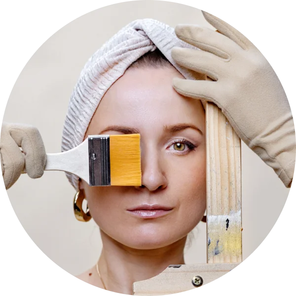 Woman with a towel on her head holding a paintbrush and a wooden stick, with a neutral background.
